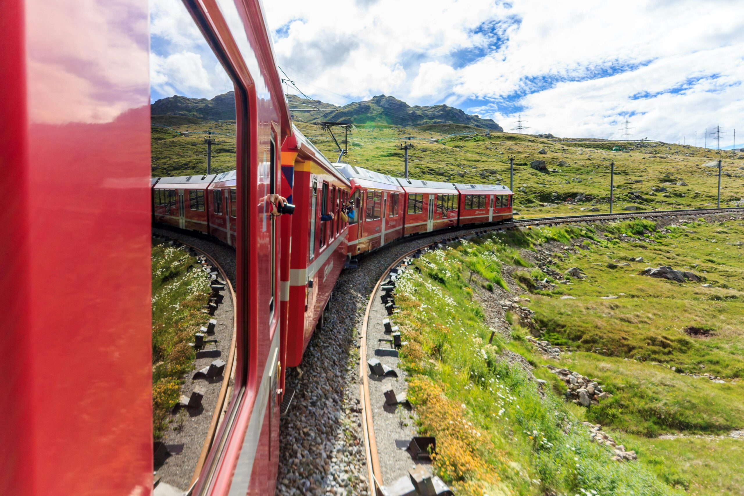 Experience a breathtaking train ride through mountainous terrain with lush greenery and vibrant red train cars.