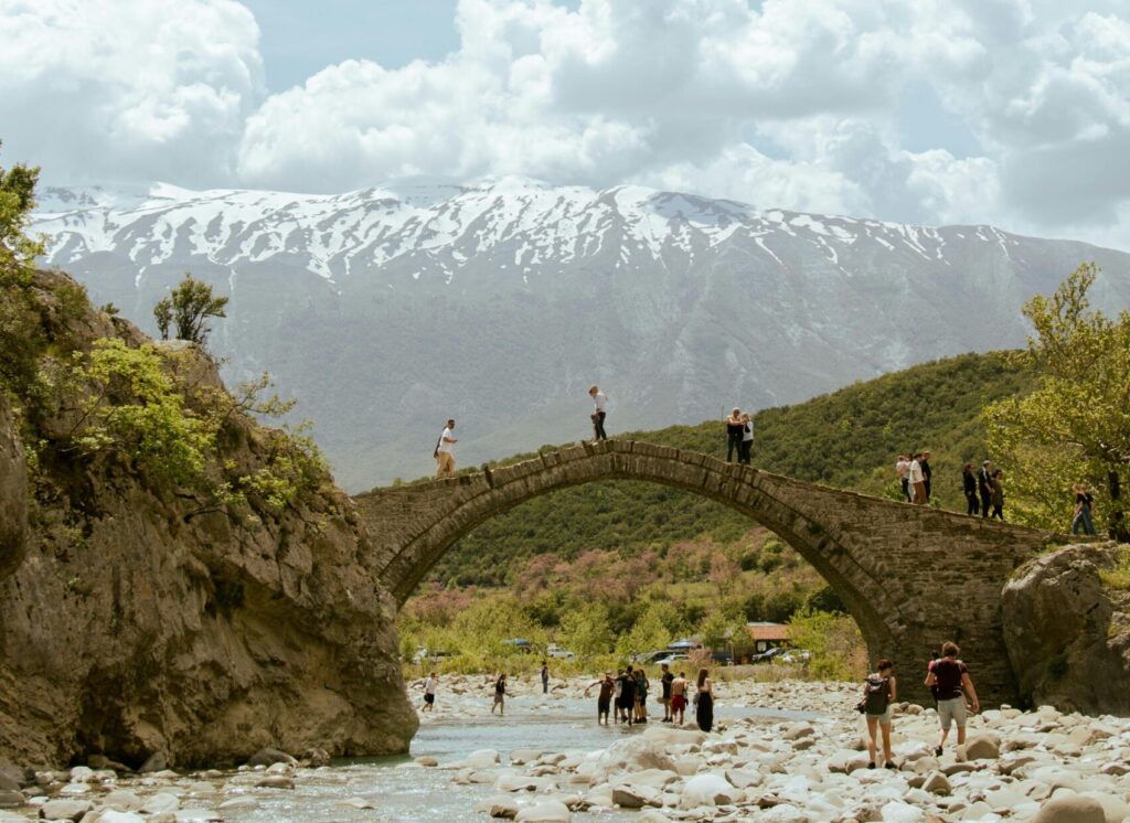Scenic view of a historic stone bridge over a river in Përmet, Albania, featuring people and mountain backdrop.