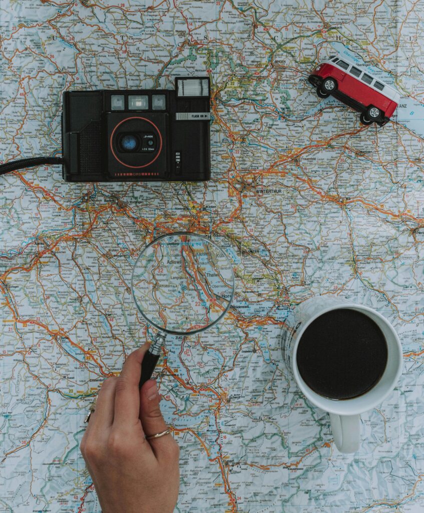 Flat lay of travel essentials: map, camera, toy van, magnifying glass, and coffee.
